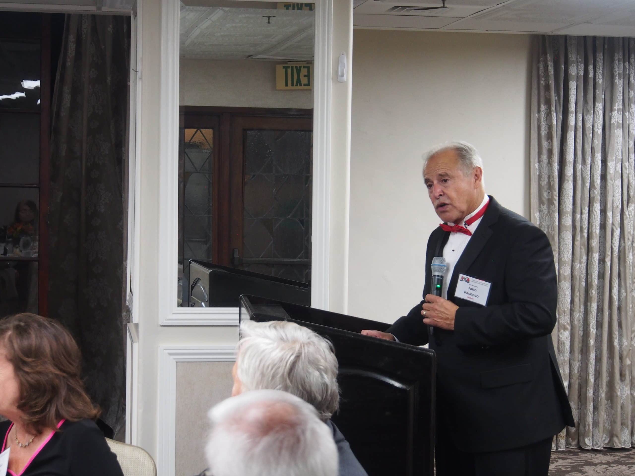 Pacheco, in a tuxedo and red bow tie, holding a microphone and standing at a podium.