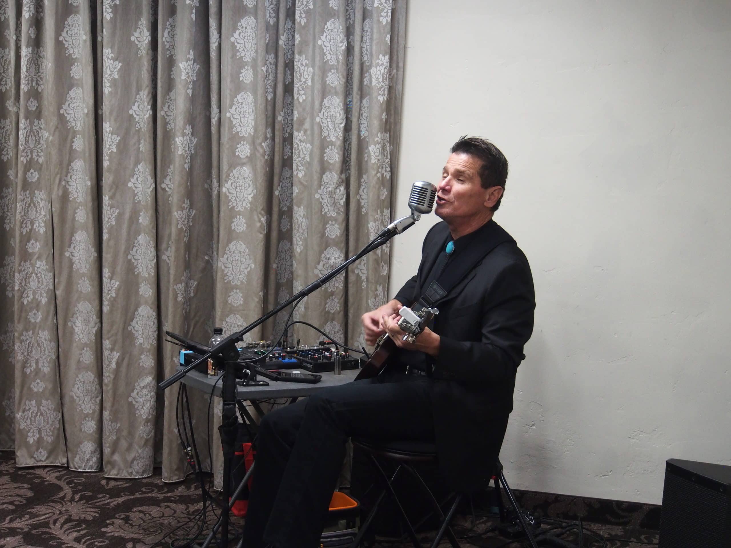 Man with short slicked hair, in black suit and black vest with bright blue tie, sings into a microphone while he strums his guitar.